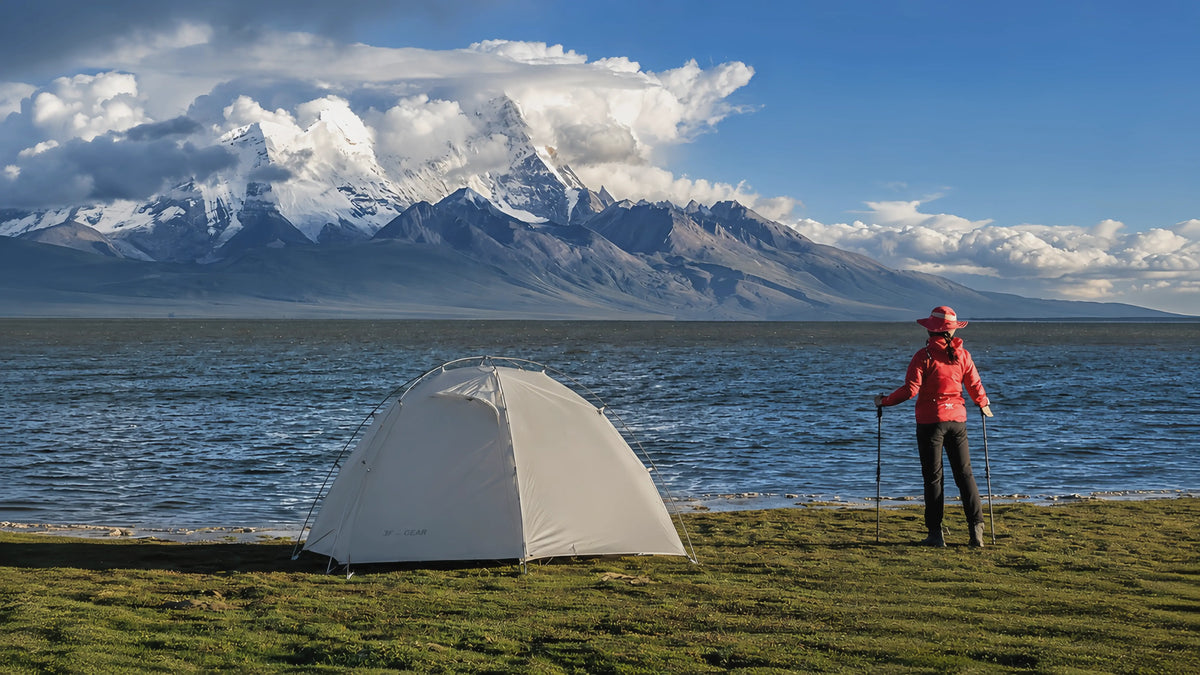 3F UL Gear Taiji 2 freestanding tent pitched beside a mountain lake, with a hiker and snow-capped peaks in the background